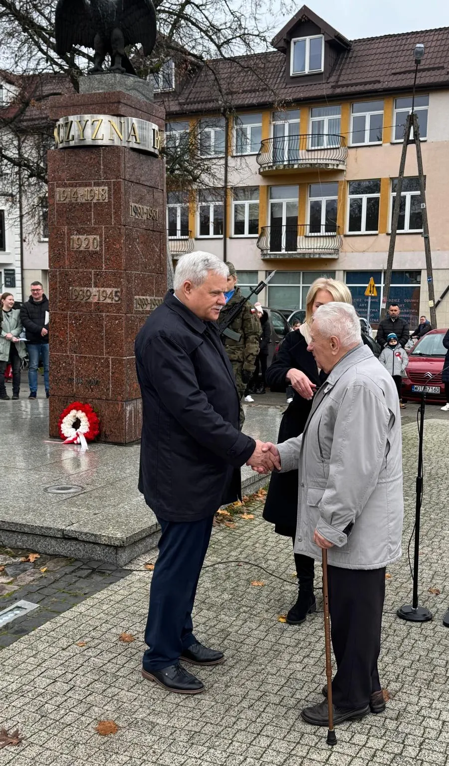 Dwóch starszych mężczyzn w kurtkach ściska sobie dłonie na zewnątrz przy pomniku z wieńcem. Obok stoi kobieta i żołnierz z bronią, w tle widoczne są budynki mieszkalne i kilka osób obserwujących scenę. Jest dzień, pora jesienna.
