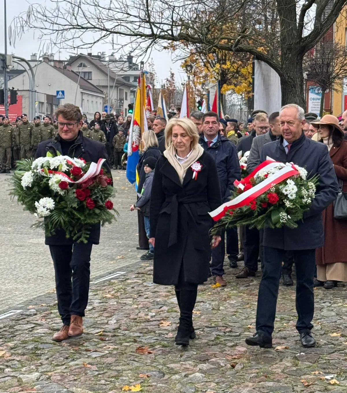 Trzy osoby niosące wieńce kwiatowe w biało-czerwonych barwach idą po kamiennej nawierzchni. Za nimi stoi grupa ludzi z flagami i żołnierze w mundurach. W tle jesienne drzewa i budynki. Dzień, na zewnątrz.
