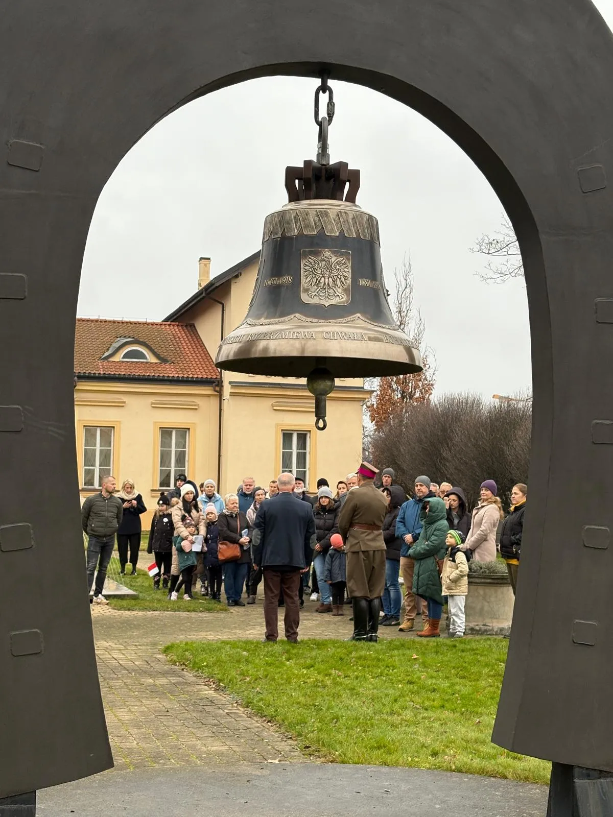 Metalowy dzwon zawieszony na dużej stalowej konstrukcji w kształcie podkowy, na tle grupy osób stojących na zewnątrz obok żółtego budynku w pochmurny dzień.