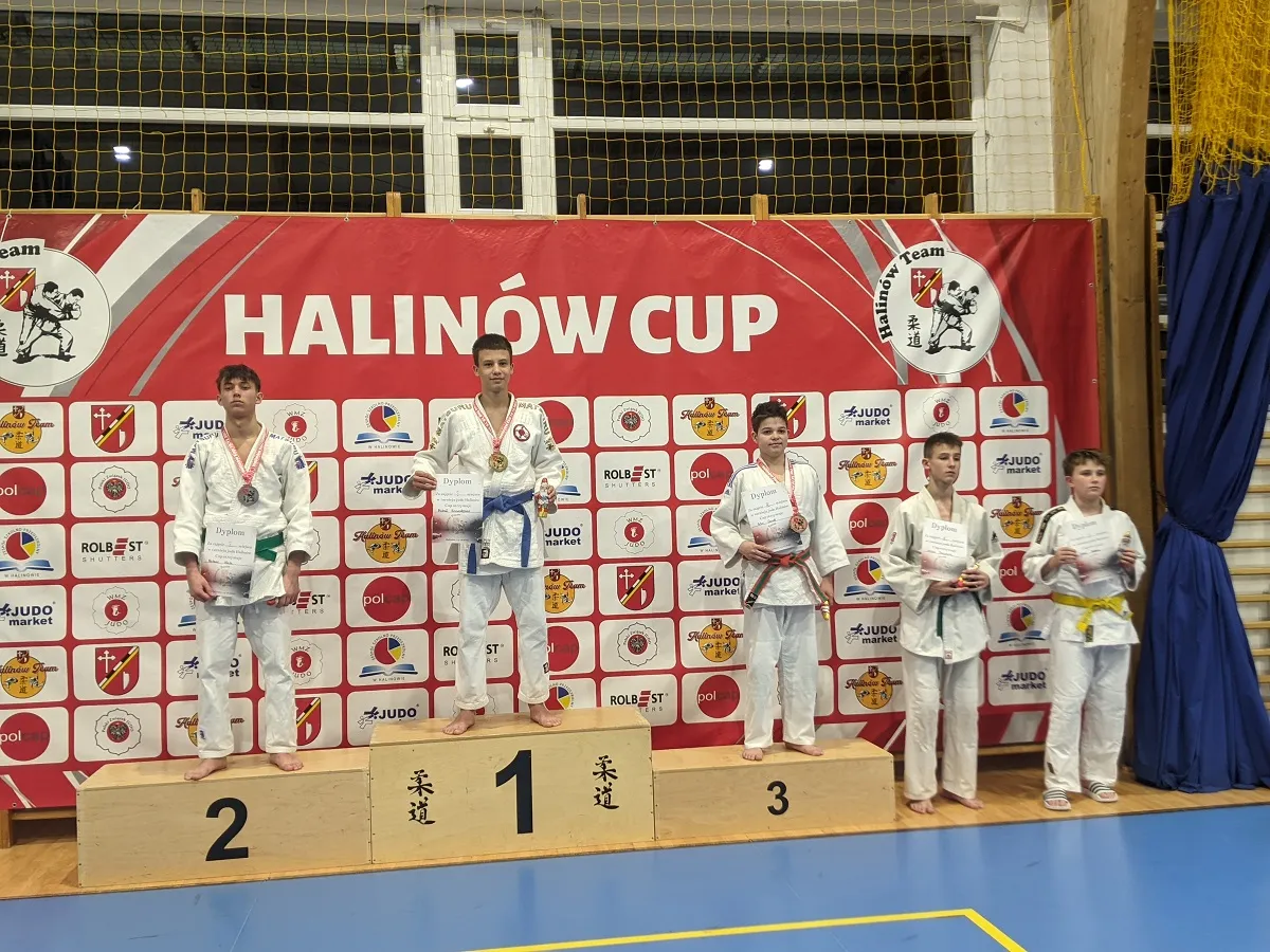 Pięciu chłopców w judogach stoi na podium i obok niego w hali sportowej z niebieską podłogą i czerwoną ścianką z napisem HALINÓW CUP oraz wieloma logotypami. Trzech chłopców stoi na podium oznaczonym numerami 1, 2 i 3, trzej trzymają dyplomy i medale. Dwaj chłopcy stoją po prawej stronie podium, również trzymają dyplomy.