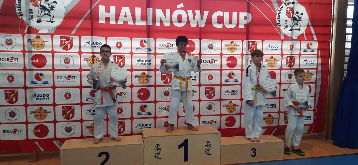 Czterech chłopców w białych judogach stoi na podium z nagrodami i dyplomami na tle czerwono-białej ścianki z logo i napisem HALINÓW CUP.