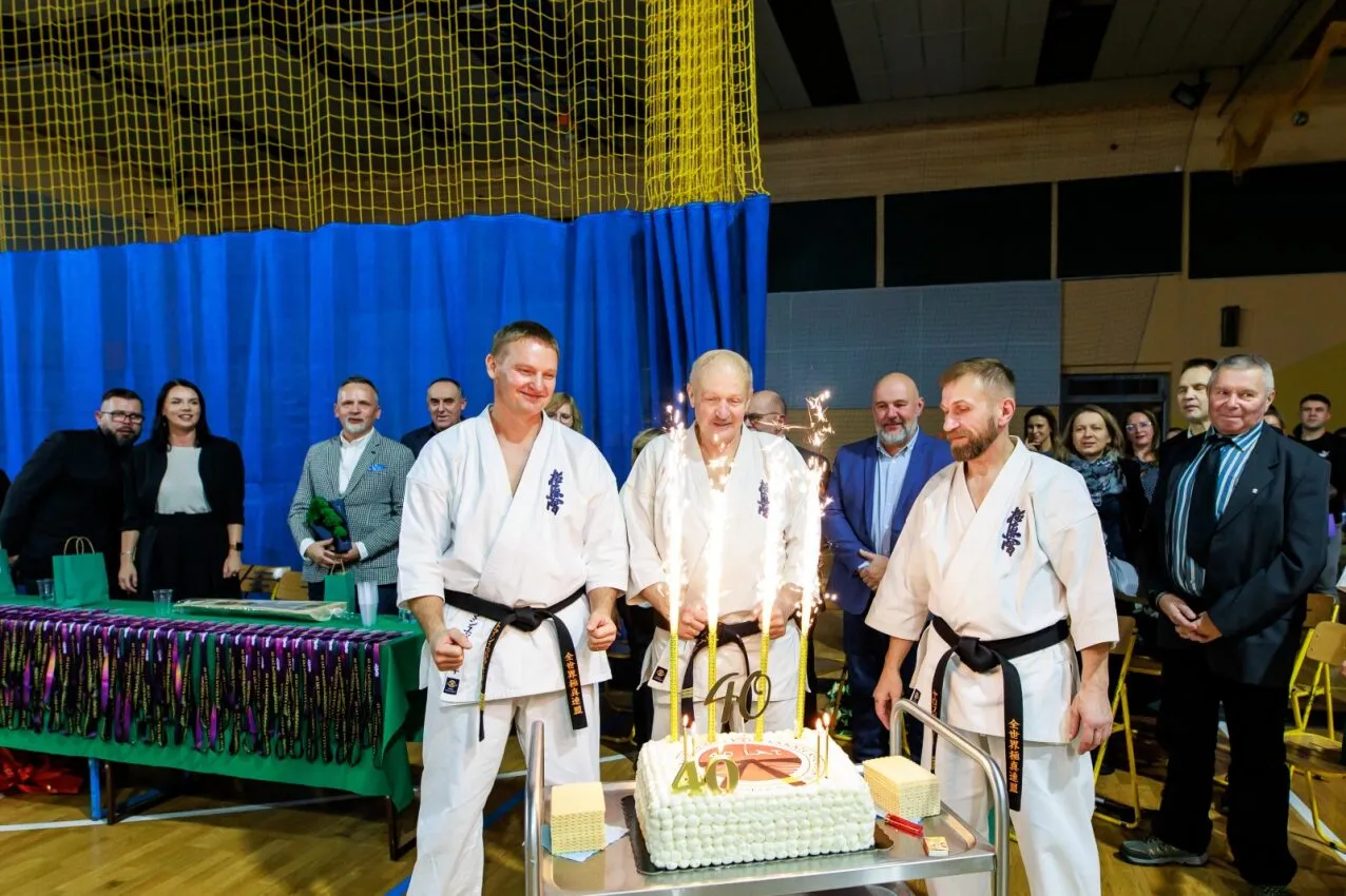 Trzech mężczyzn w białych kimonach z czarnymi pasami stoi przed dużym tortem z zapalonymi świeczkami. W tle grupa osób obserwuje uroczystość w sali sportowej z niebieską zasłoną i żółtą siatką. Na stole obok leżą liczne kolorowe pasy i dyplomy.