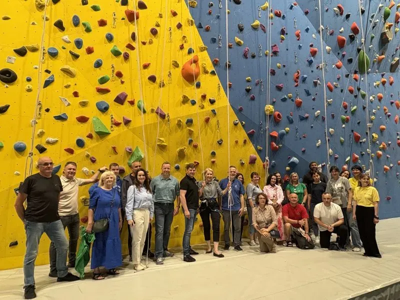 Grupa ludzi stoi na ścianie do wspinaczki z kolorowymi chwytami na żółtej i niebieskiej powierzchni.