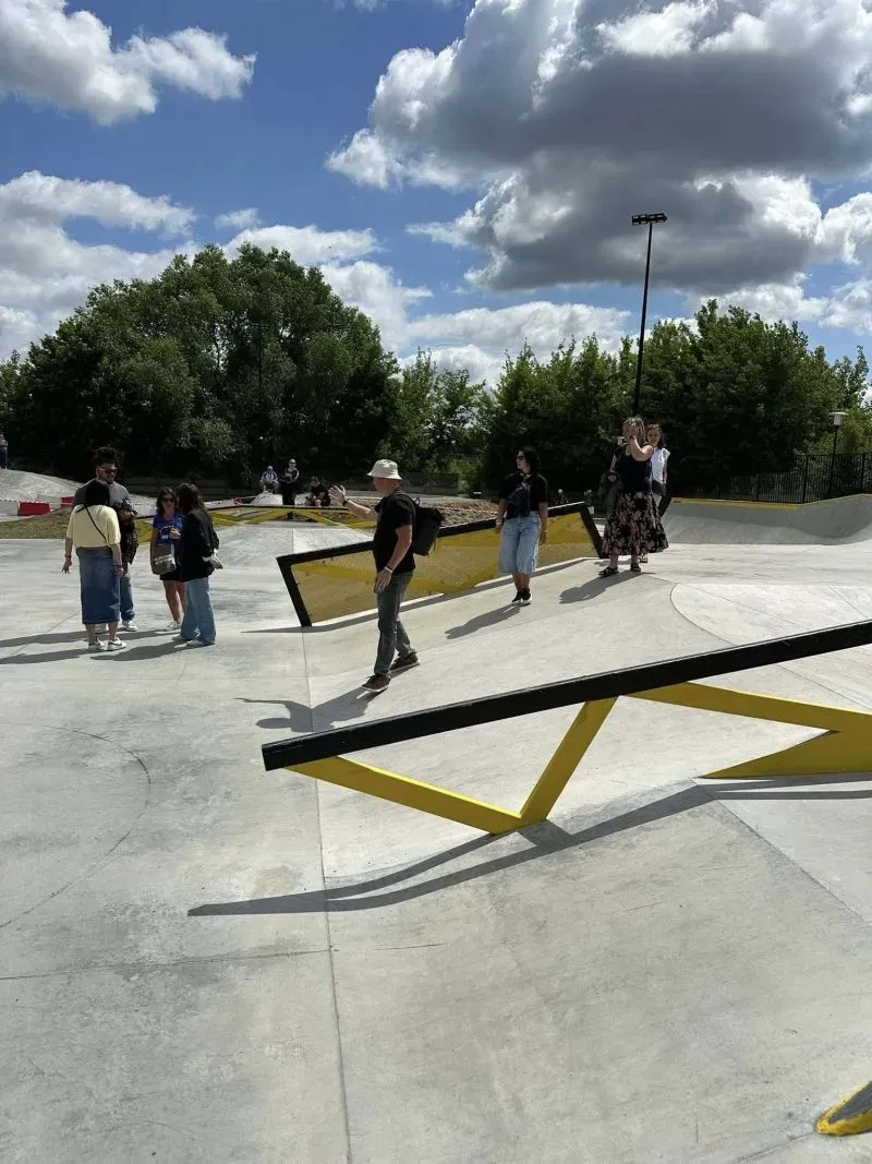 Betonowy skatepark z metalowymi przeszkodami i kilkoma osobami spacerującymi po nim.