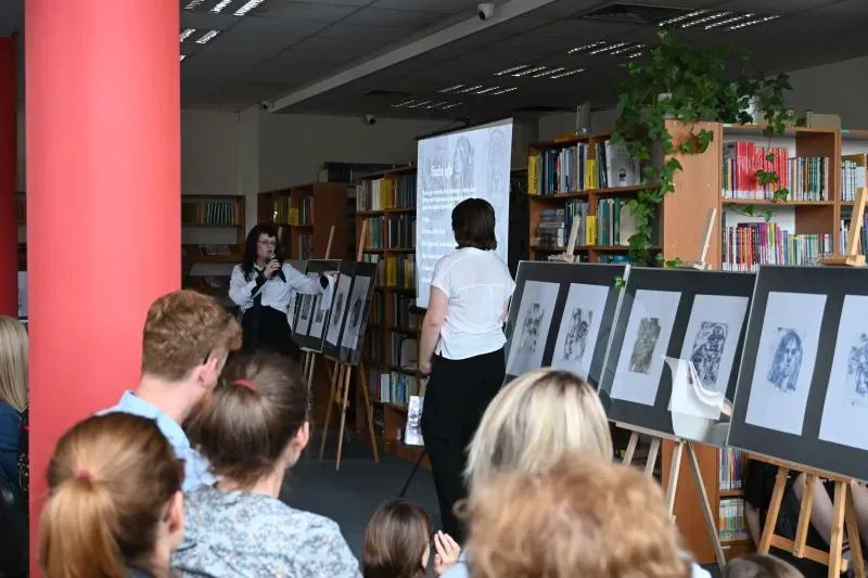 Grupa ludzi słuchających kobiety z mikrofonem prezentującej serię czarno-białych rysunków na sztalugach w bibliotece.