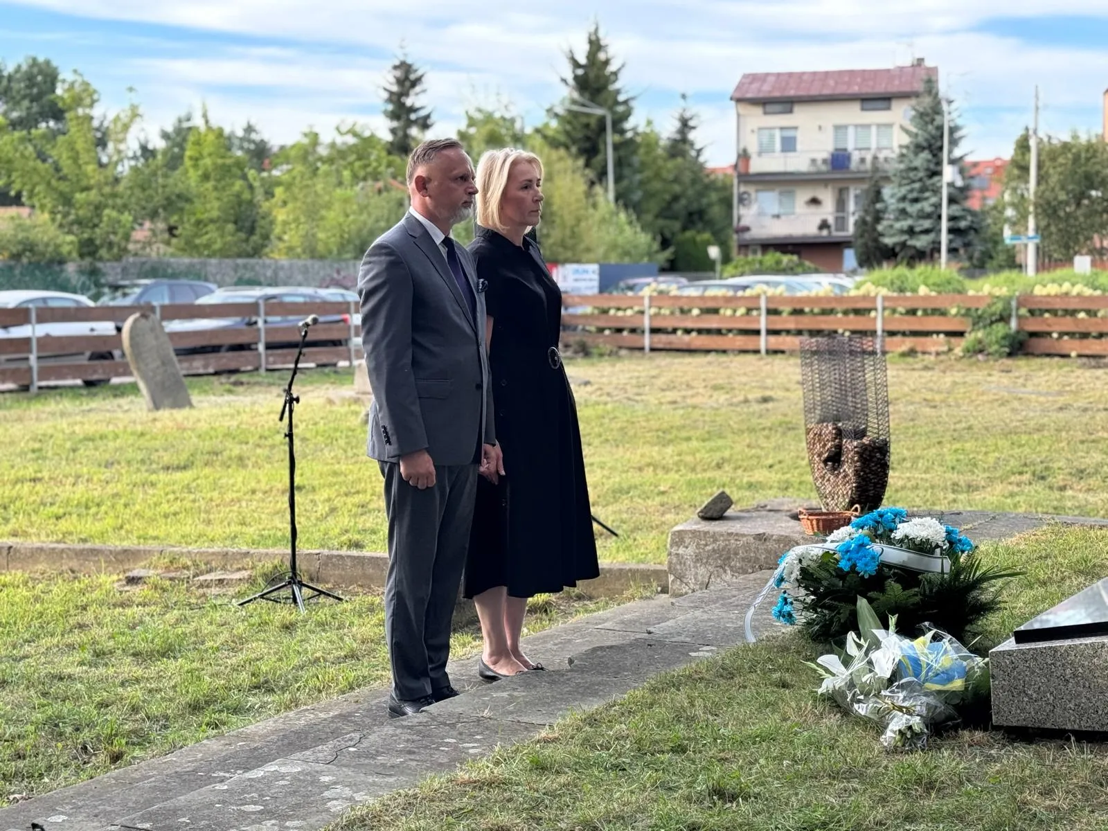 Dwóch dorosłych stoi obok siebie na zewnątrz na skraju trawnika, obok nich leżą wieńce kwiatowe i znajduje się metalowa rzeźba.