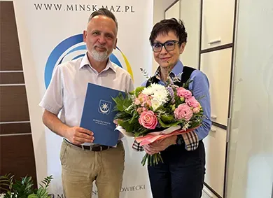 Mężczyzna w jasnej koszuli i spodniach trzyma teczkę z napisem „Burmistrz Miasta”, obok stoi kobieta z krótkimi włosami i okularami, ubrana w niebieską koszulę i ciemne spodnie, trzyma bukiet kwiatów.