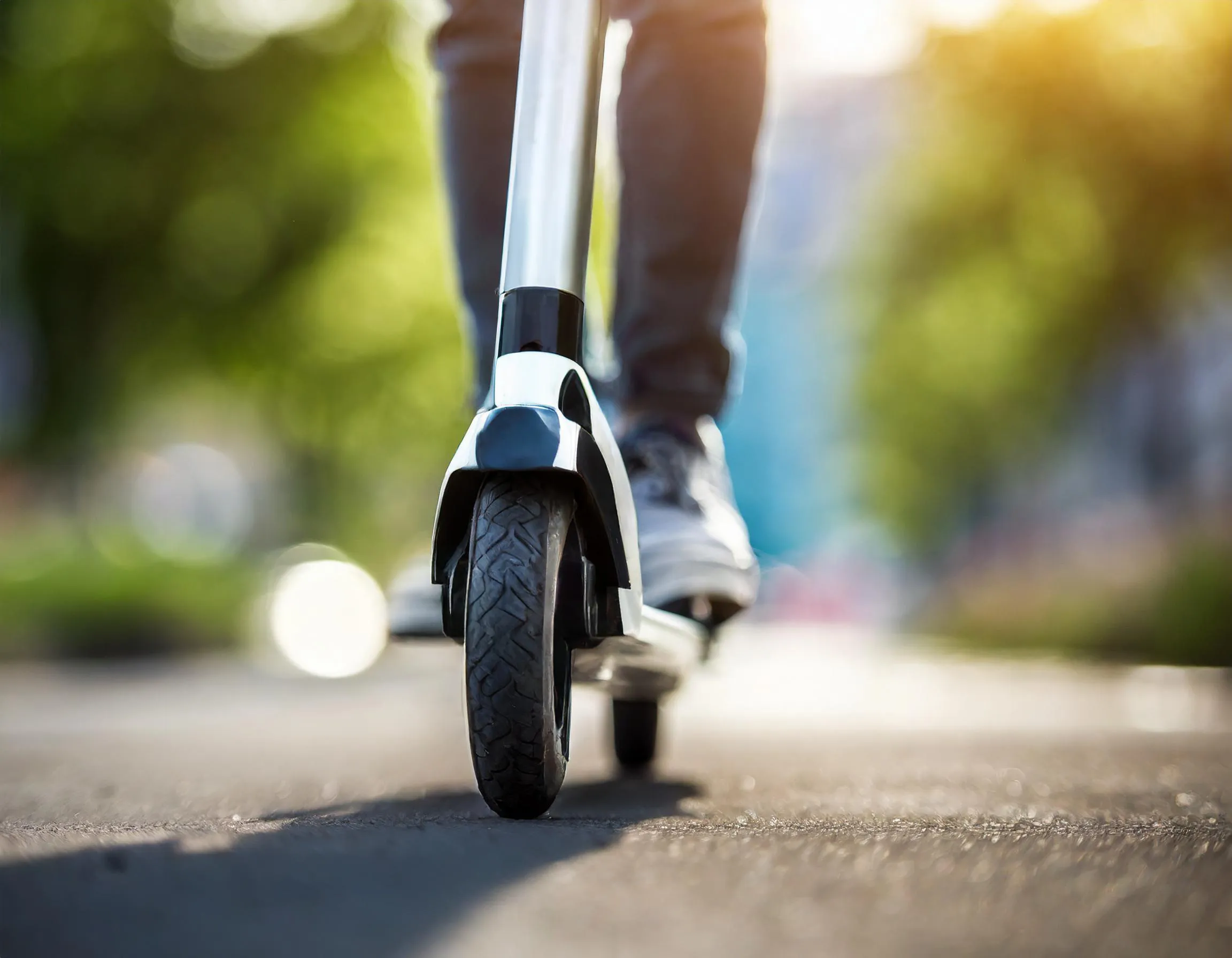 Biała hulajnoga elektryczna z przodu, na której stoi osoba w ciemnych spodniach i białych butach na asfaltowej nawierzchni.
