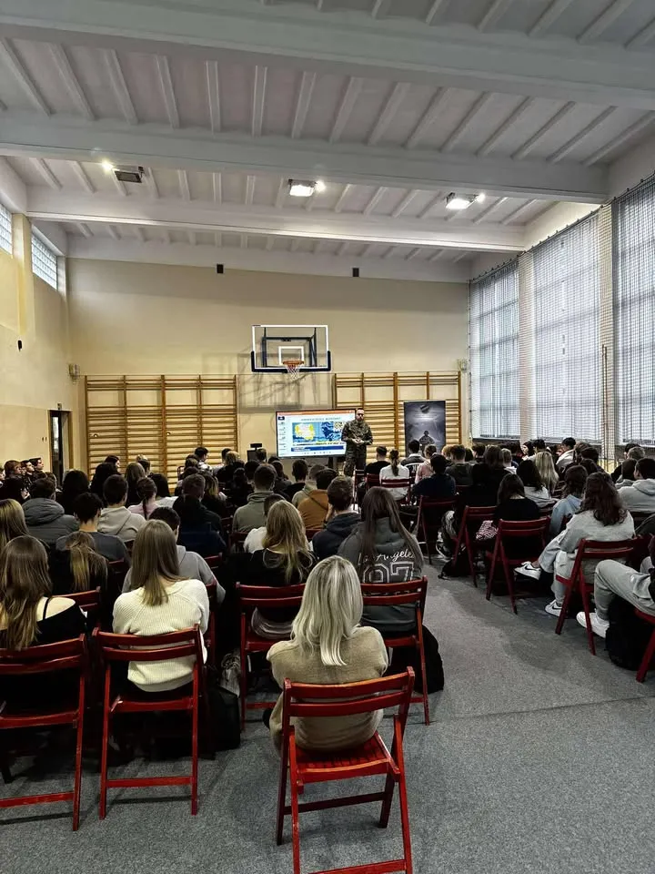 W sali gimnastycznej w ciągu dnia kilkudziesięciu młodych ludzi siedzi na czerwonych krzesłach i słucha wykładu prowadzonego przez osobę stojącą przed tablicą interaktywną. Ściany są jasne, obok tablicy znajduje się kosz do koszykówki, a po prawej stronie duże okna z kratami.