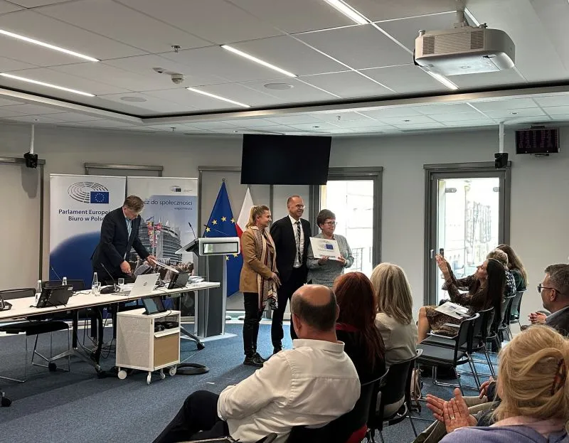 Grupa osób stoi przy mównicy z flagami Unii Europejskiej i Polski, podczas gdy publiczność siedzi i klaska.