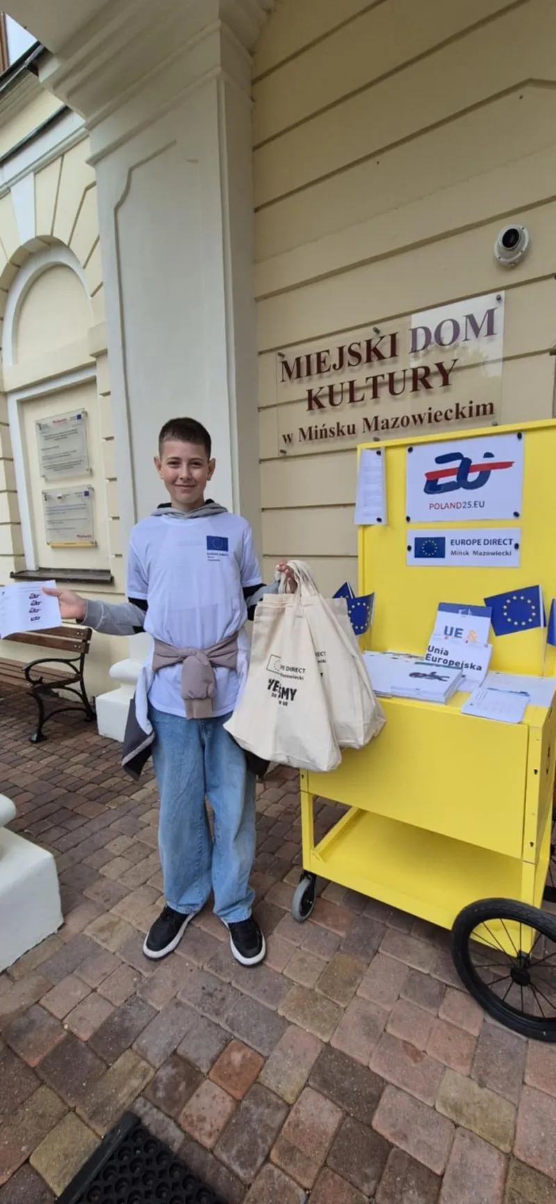 Chłopiec w białej koszulce stoi obok żółtego wózka z materiałami promocyjnymi przed budynkiem Miejskiego Domu Kultury w Mińsku Mazowieckim.