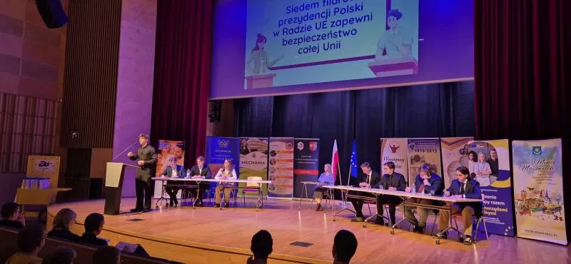 Na scenie w sali konferencyjnej siedzi dziewięć osób przy dwóch stołach ustawionych na przeciw siebie, a jedna osoba stoi przy mównicy i przemawia, w tle widoczny jest ekran z polskim tekstem oraz flagi Polski i Unii Europejskiej.