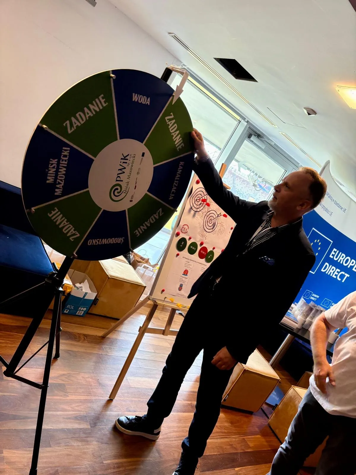 Mężczyzna w garniturze obraca koło fortuny ustawione na stojaku w jasnym pomieszczeniu z drewnianą podłogą. W tle tablica na stojaku i niebieski baner z napisem Europe Direct. Obok stoi druga osoba w białej koszulce.