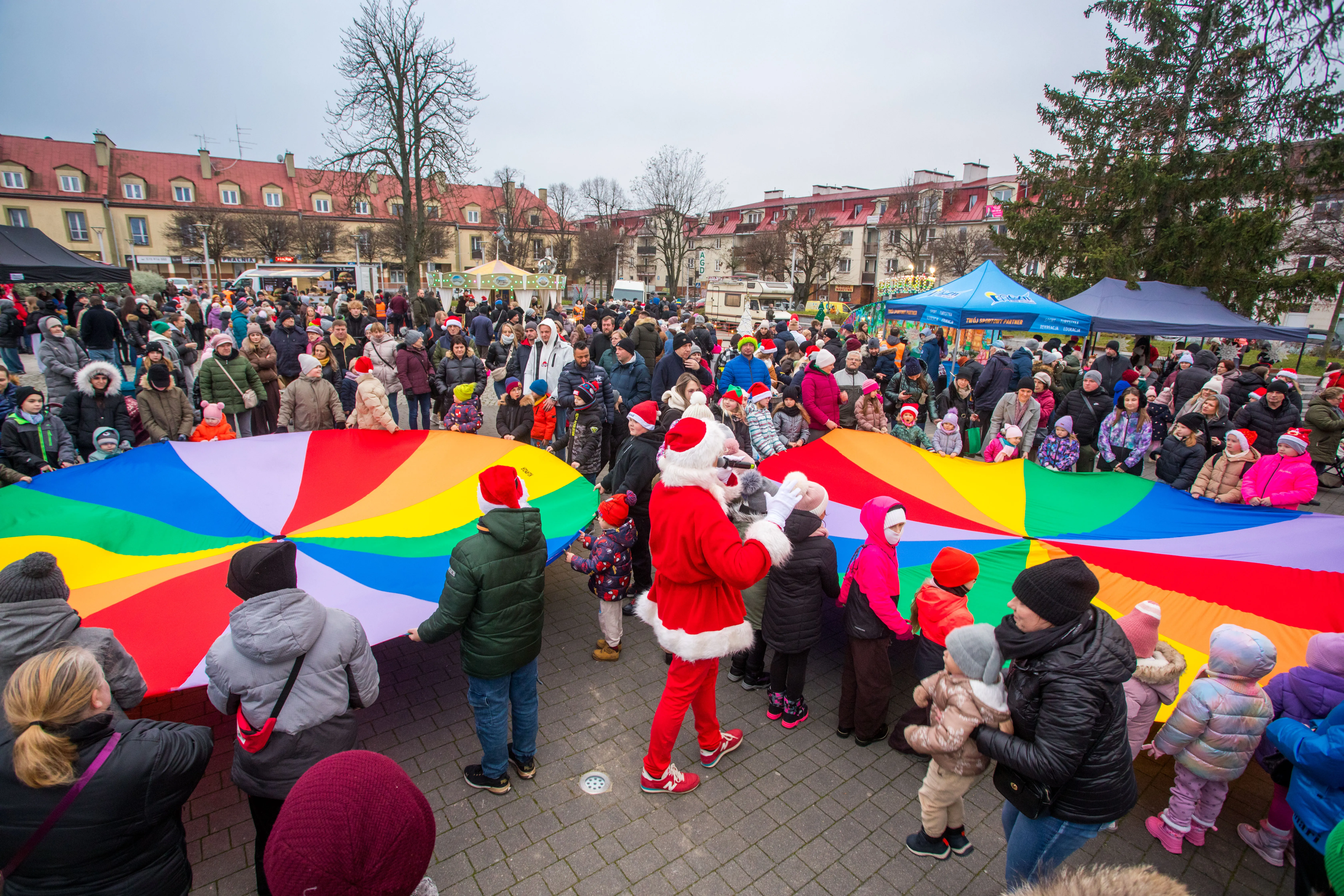 Duża grupa ludzi, w tym dzieci i dorosłych, stoi na zewnątrz na placu miejskim w chłodny dzień. W centrum trzy osoby trzymają razem duży, kolorowy, okrągły materiał w jaskrawych kolorach tęczy. Jedna osoba ubrana jak Mikołaj z czerwonym strojem i białym futrem stoi obok. W tle budynki mieszkalne oraz namioty i tłum ludzi.