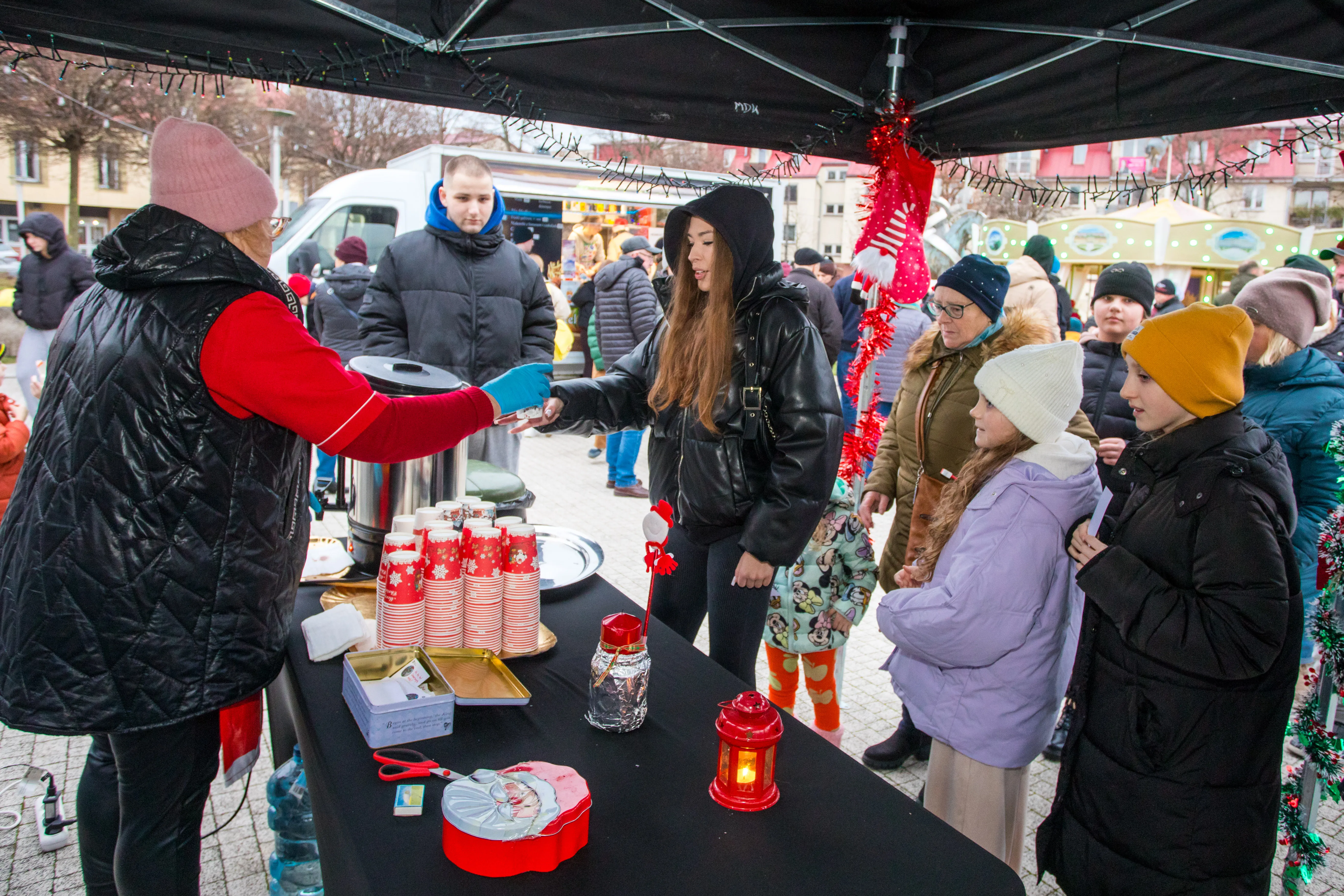 Stoisko na zewnątrz pod czarnym namiotem, kilka osób w zimowych ubraniach stoi w kolejce po napoje w papierowych kubkach z czerwonym świątecznym wzorem. Kobieta w różowej czapce i kamizelce podaje napój młodej kobiecie w czarnej kurtce z kapturem. Na stole są dekoracje świąteczne, w tym czerwony lampion i pudełko z Mikołajem. W tle widać więcej ludzi oraz budynki.