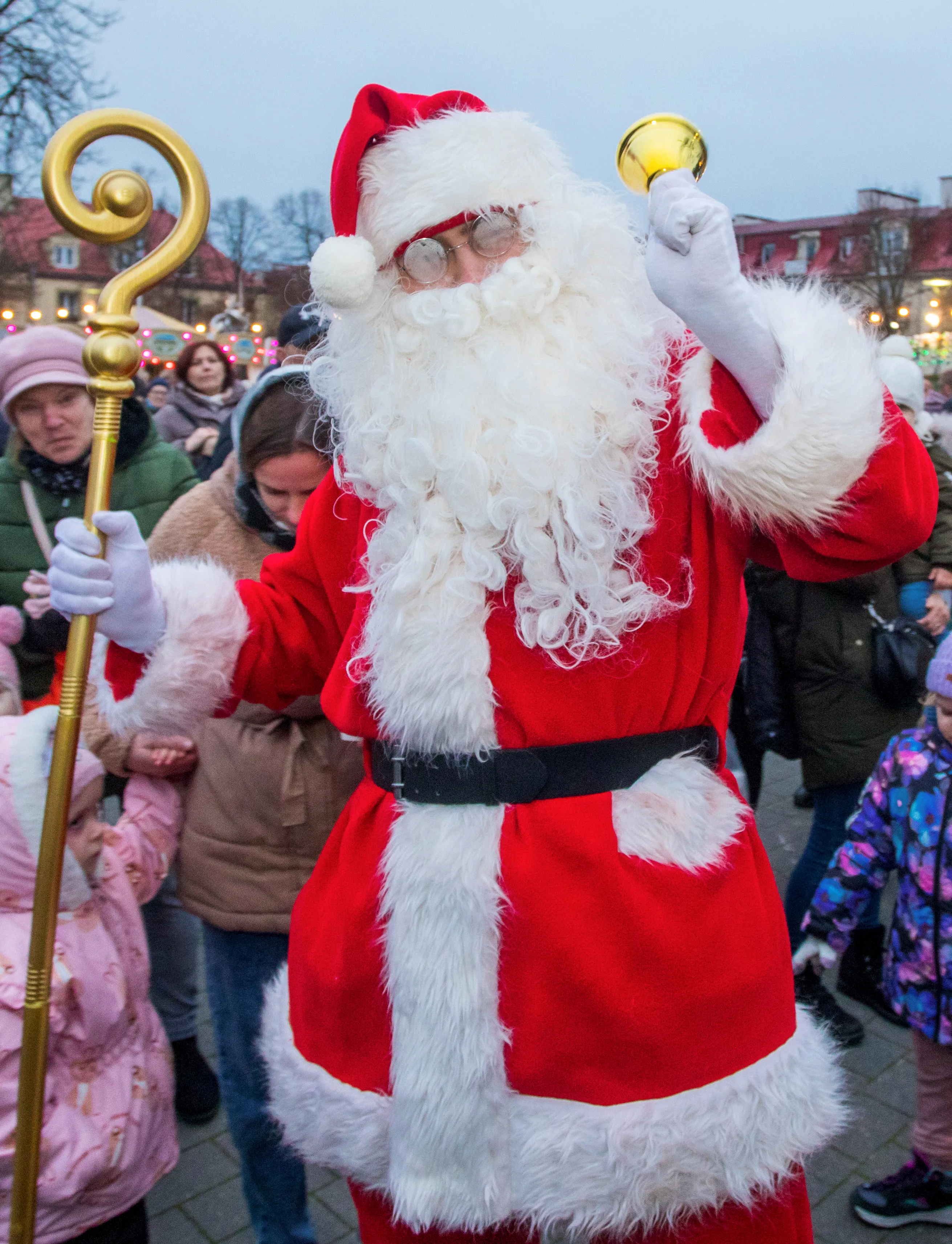 Mężczyzna przebrany za Mikołaja w czerwonym stroju z białym futerkiem, trzyma złoty berło i dzwonek, stoi na zewnątrz wśród ludzi w zimowych ubraniach. W tle budynki i oświetlenie.