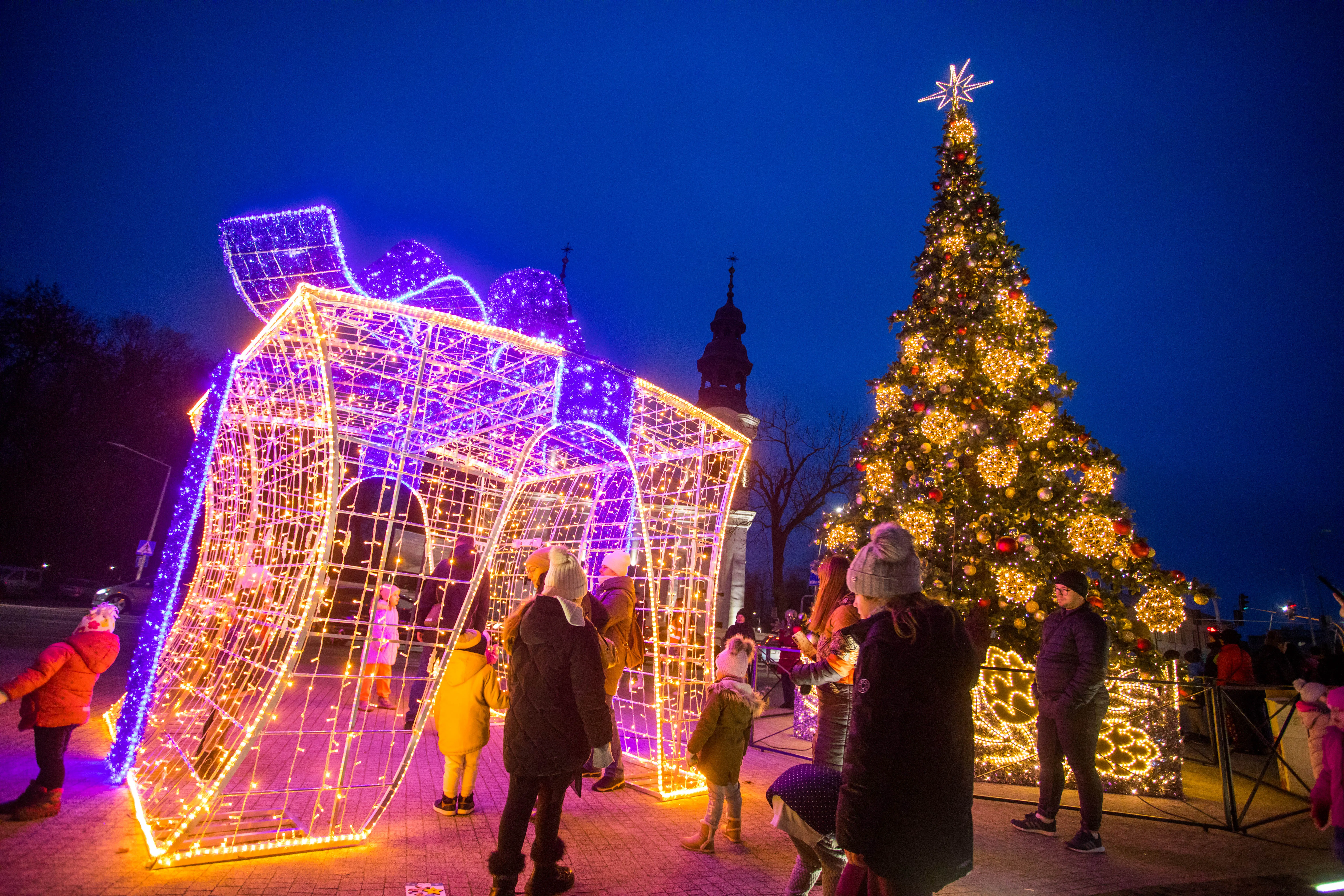 Grupa osób, w tym dzieci i dorośli, stoi obok dużej konstrukcji świetlnej w kształcie prezentu i wysokiej, oświetlonej choinki z gwiazdą na szczycie, na zewnątrz nocą. W tle widać zabytkową wieżę kościoła.