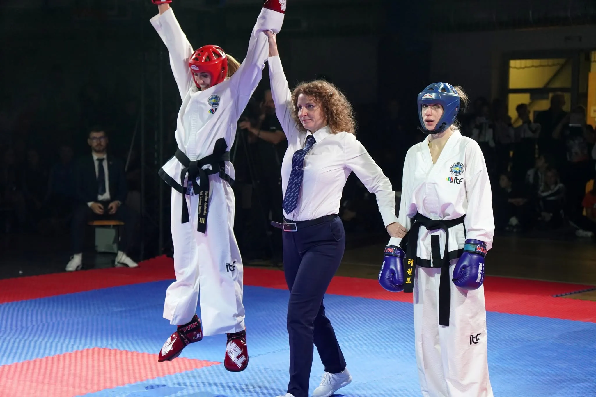 Sędzina podnosi rękę zawodniczki w czerwonym kasku, która skacze z radości, obok stoi zawodniczka w niebieskim kasku na niebiesko-czerwonej macie w hali sportowej.