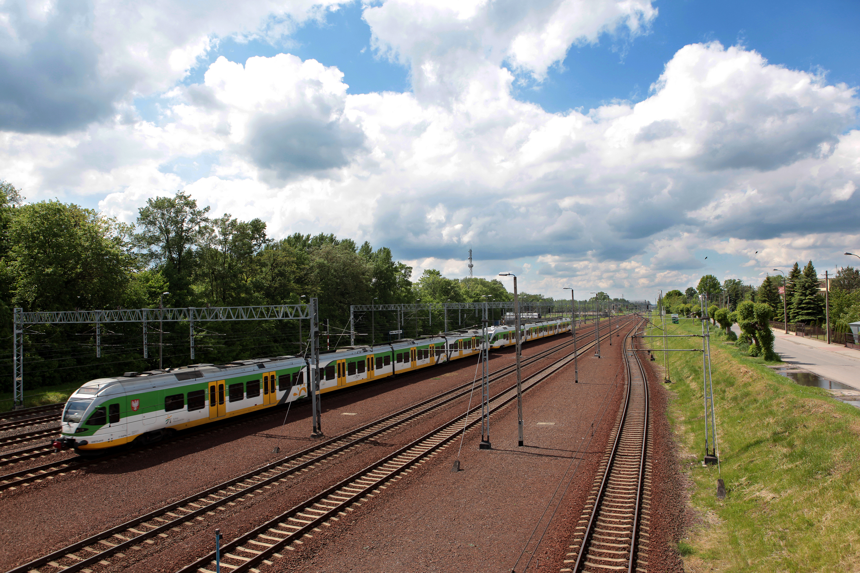 Pociąg osobowy w kolorach białym, zielonym i żółtym na torach kolejowych obok zielonych drzew i drogi.