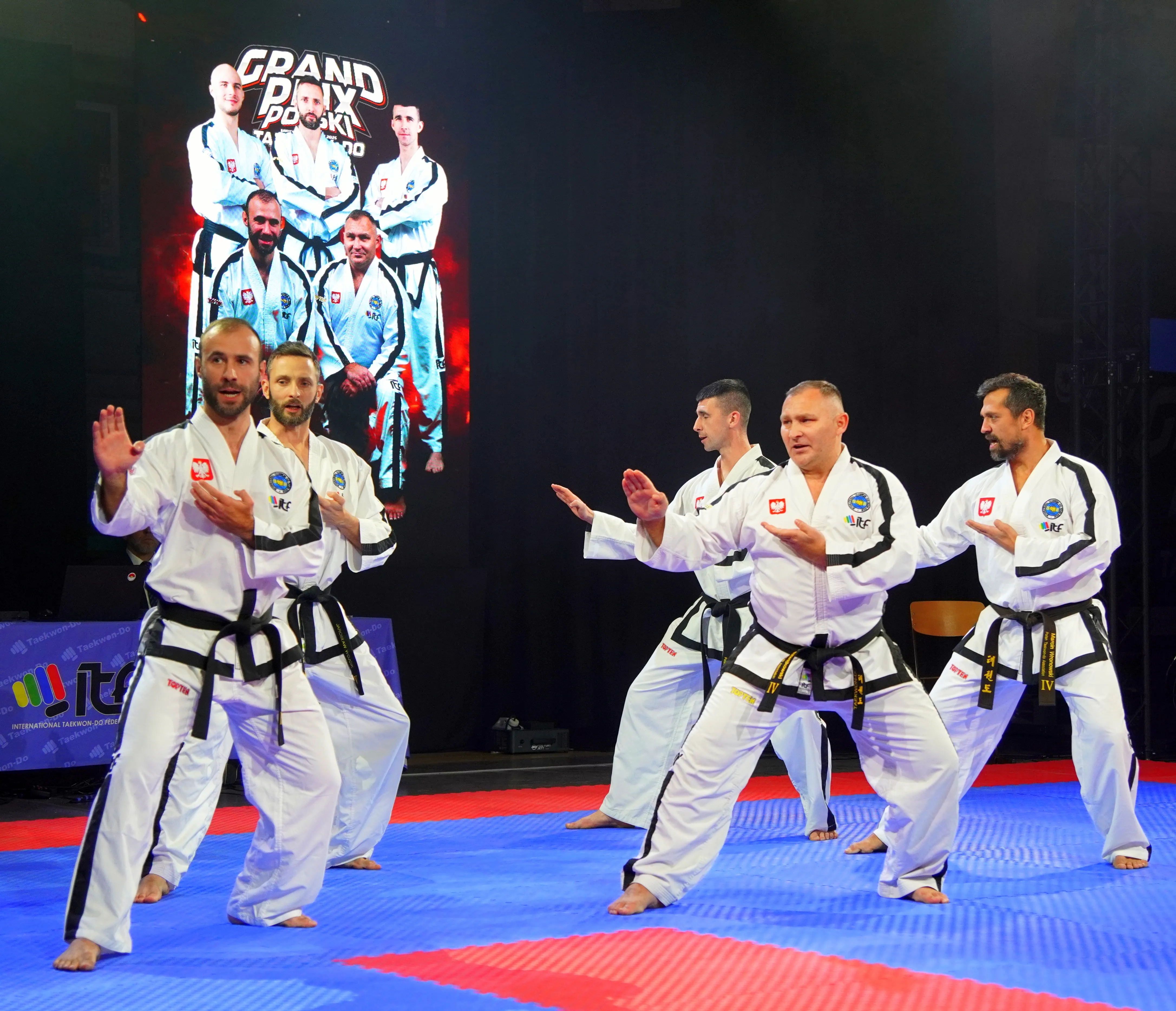 Pięciu mężczyzn w białych strojach do taekwondo z czarnymi pasami wykonuje ćwiczenia na niebiesko-czerwonej macie na ciemnej scenie, z dużym ekranem z tyłu pokazującym grupowe zdjęcie tych samych mężczyzn w strojach.