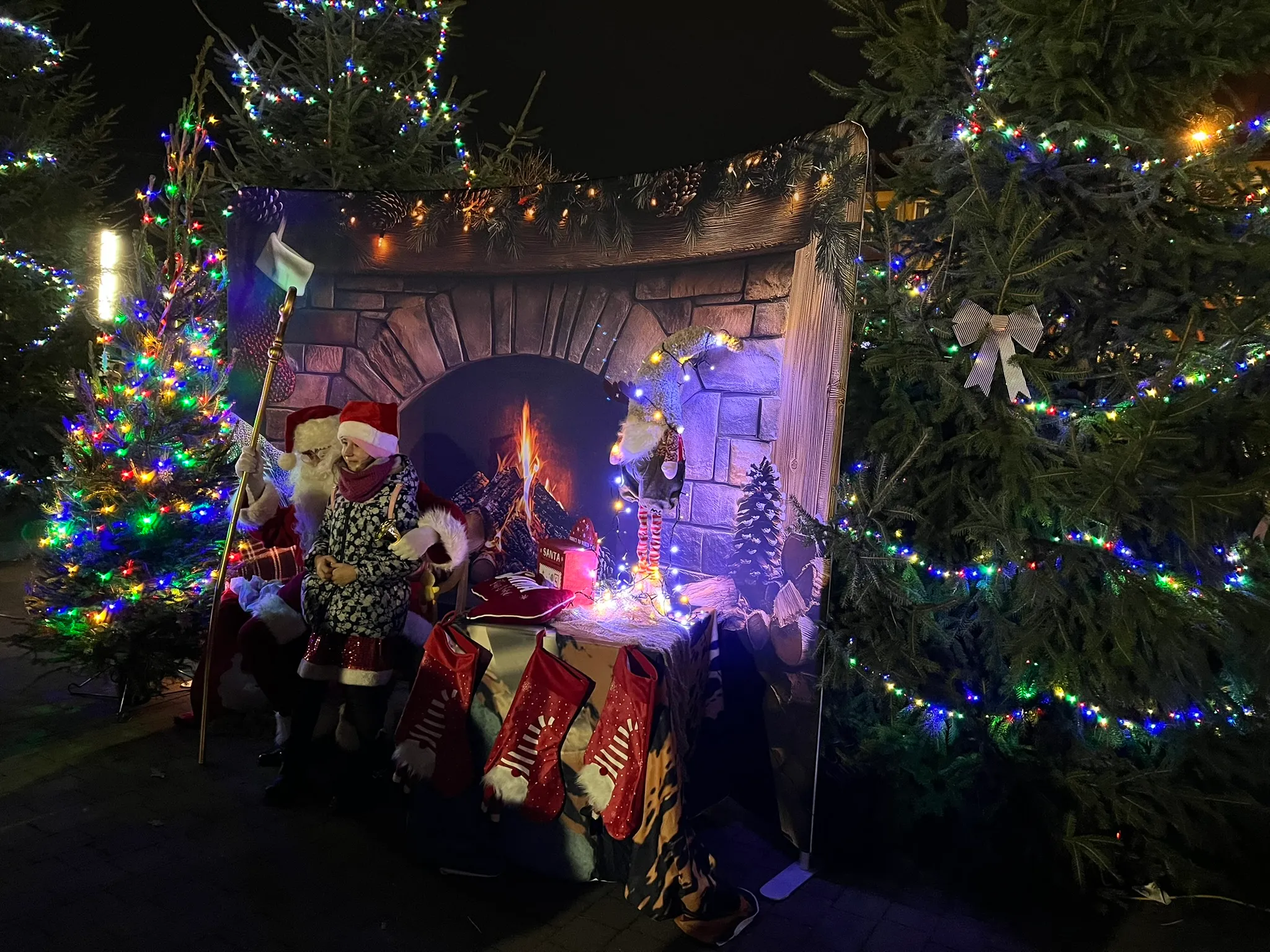 Dzień lub noc na zewnątrz, kobieta w czerwonej czapce świętego Mikołaja siedzi obok mężczyzny w stroju Mikołaja przed dekoracją z motywem kominka, otoczeni choinkami udekorowanymi kolorowymi światełkami.