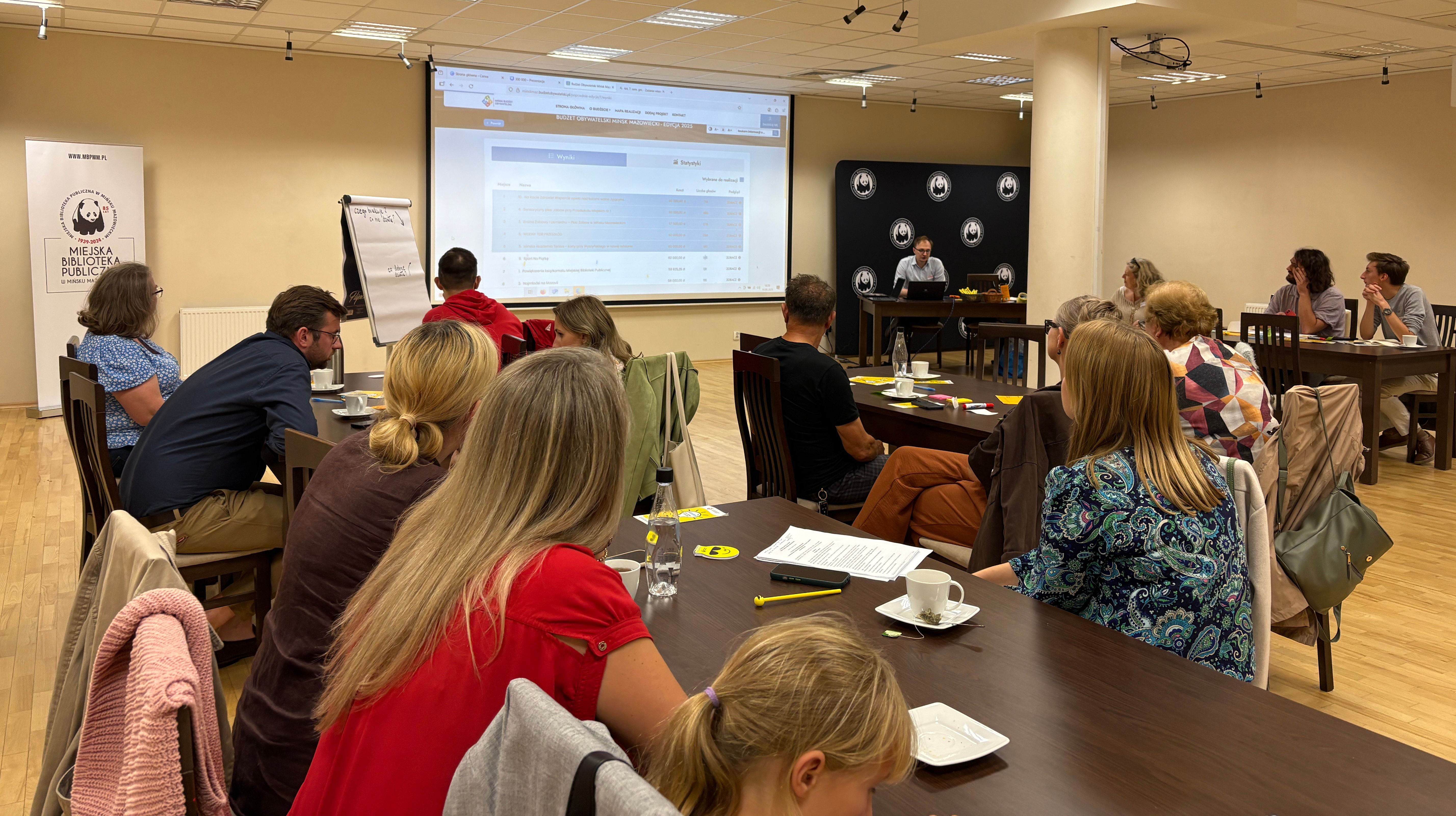 Kilkunastu dorosłych i jedno dziecko siedzi przy stołach w sali konferencyjnej, słuchając prezentera przy komputerze przed dużym ekranem z wyświetlonym tekstem.