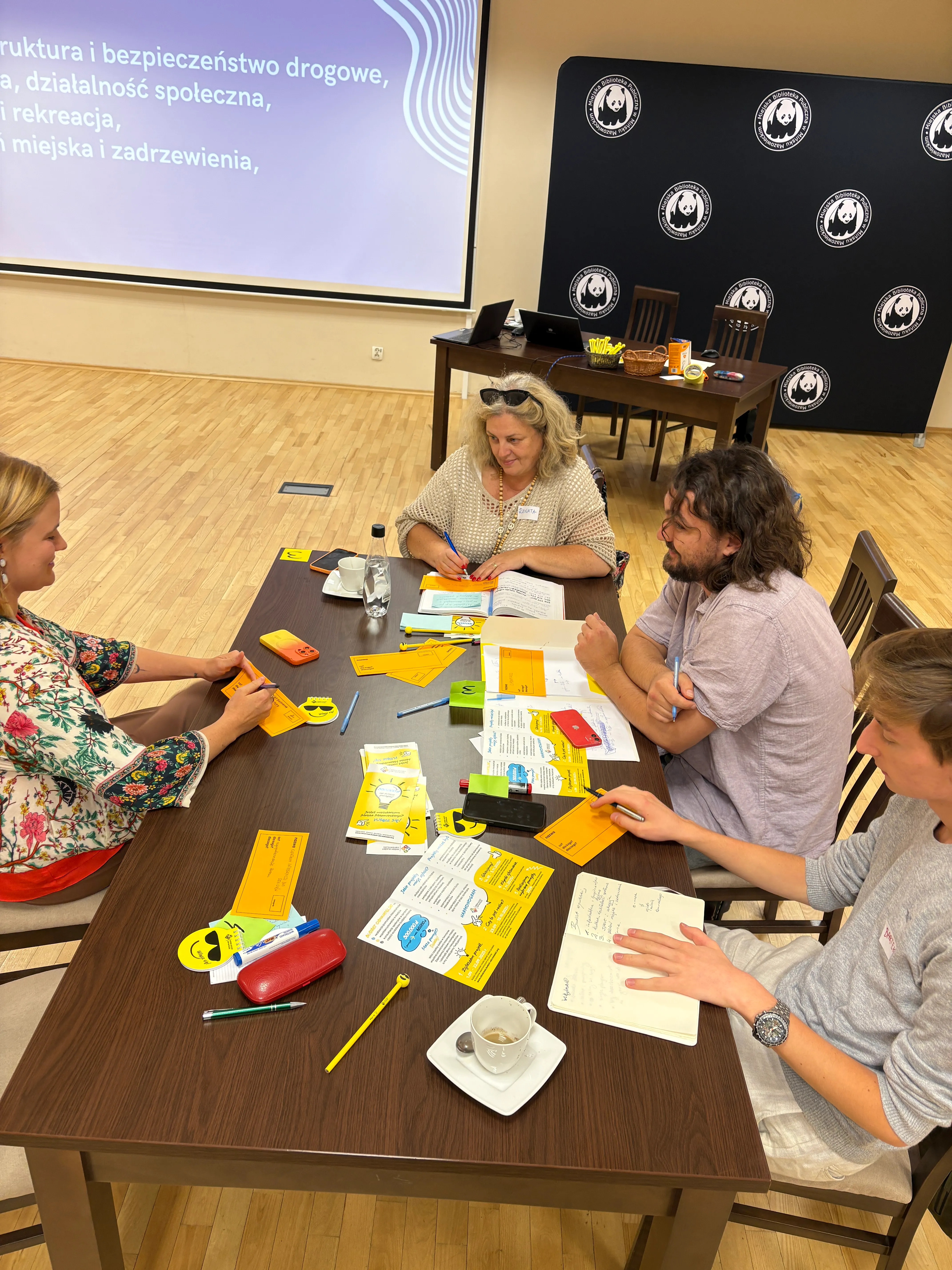 Cztery osoby siedzą wokół stołu z notatkami, kartkami i długopisami podczas warsztatów w sali z drewnianą podłogą, ekranem z prezentacją i ścianką z logo.