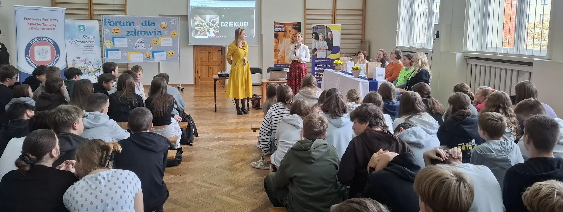 Uczniowie siedzą na sali gimnastycznej i słuchają dwóch kobiet prowadzących prelekcję. Kobiety stoją przy stole z dekoracjami i plakatach. Za nimi widać planszę oraz ekran z napisem „Dziękuję!”. W pomieszczeniu duże okna wpuszczające światło dzienne.