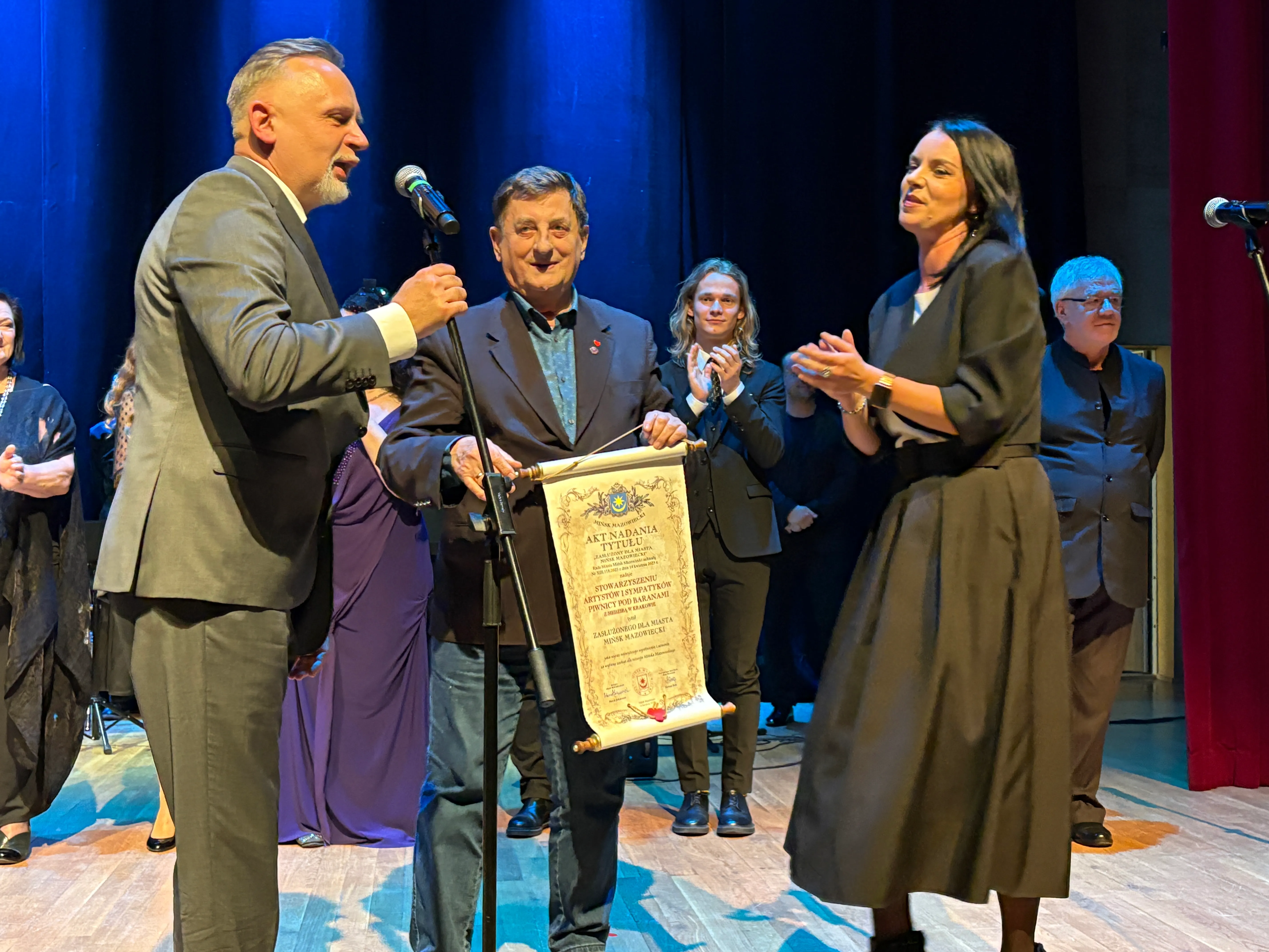 Na scenie w sali teatralnej stoi kilku ludzi. Mężczyzna po lewej w garniturze mówi do mikrofonu. Starszy mężczyzna obok trzyma zwój z tekstem. Kobieta po prawej klaska. W tle stoją inni ludzie w eleganckich strojach, oświetleni na tle ciemnej zasłony.