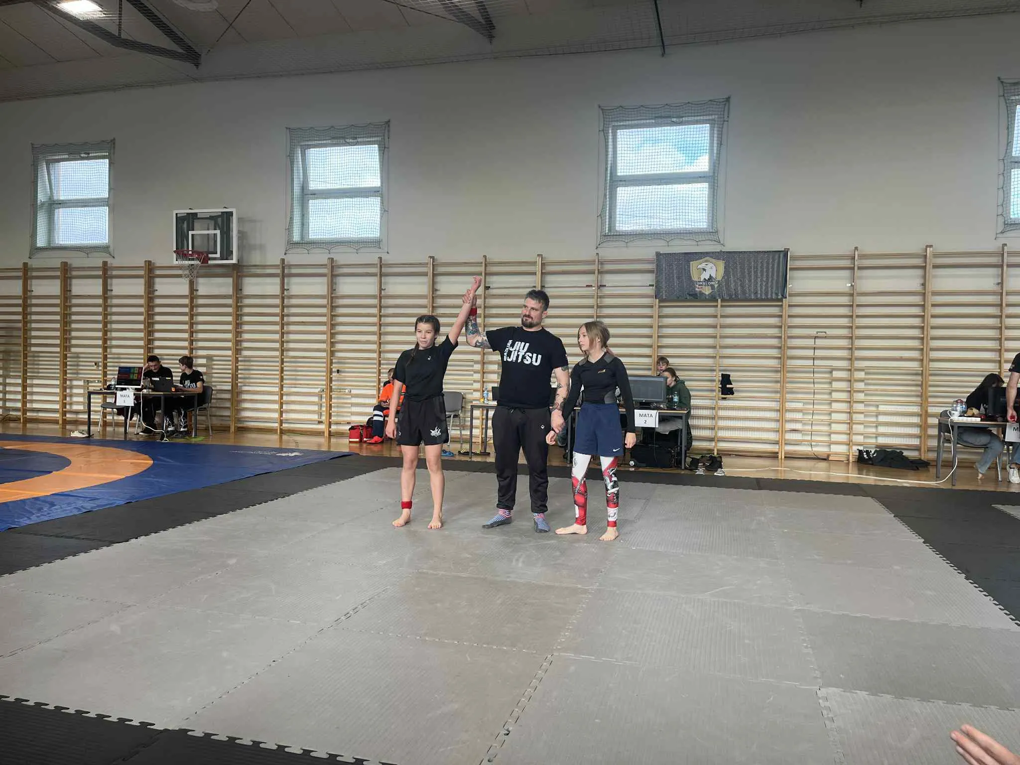 W hali sportowej na macie do walki stoi trójka osób: sędzia podnosi w górę rękę dziewczynki stojącej po lewej, druga dziewczynka stoi po prawej stronie. W tle widać ławki i osoby siedzące przy stolikach oraz drabinki gimnastyczne na ścianie.