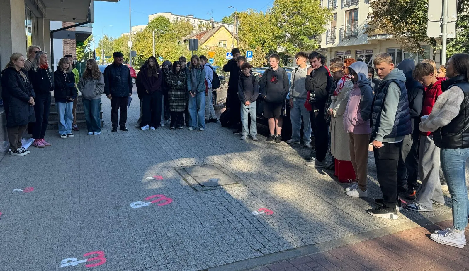 Grupa około trzydziestu osób stoi w skupieniu na chodniku przy ulicy w dzień.