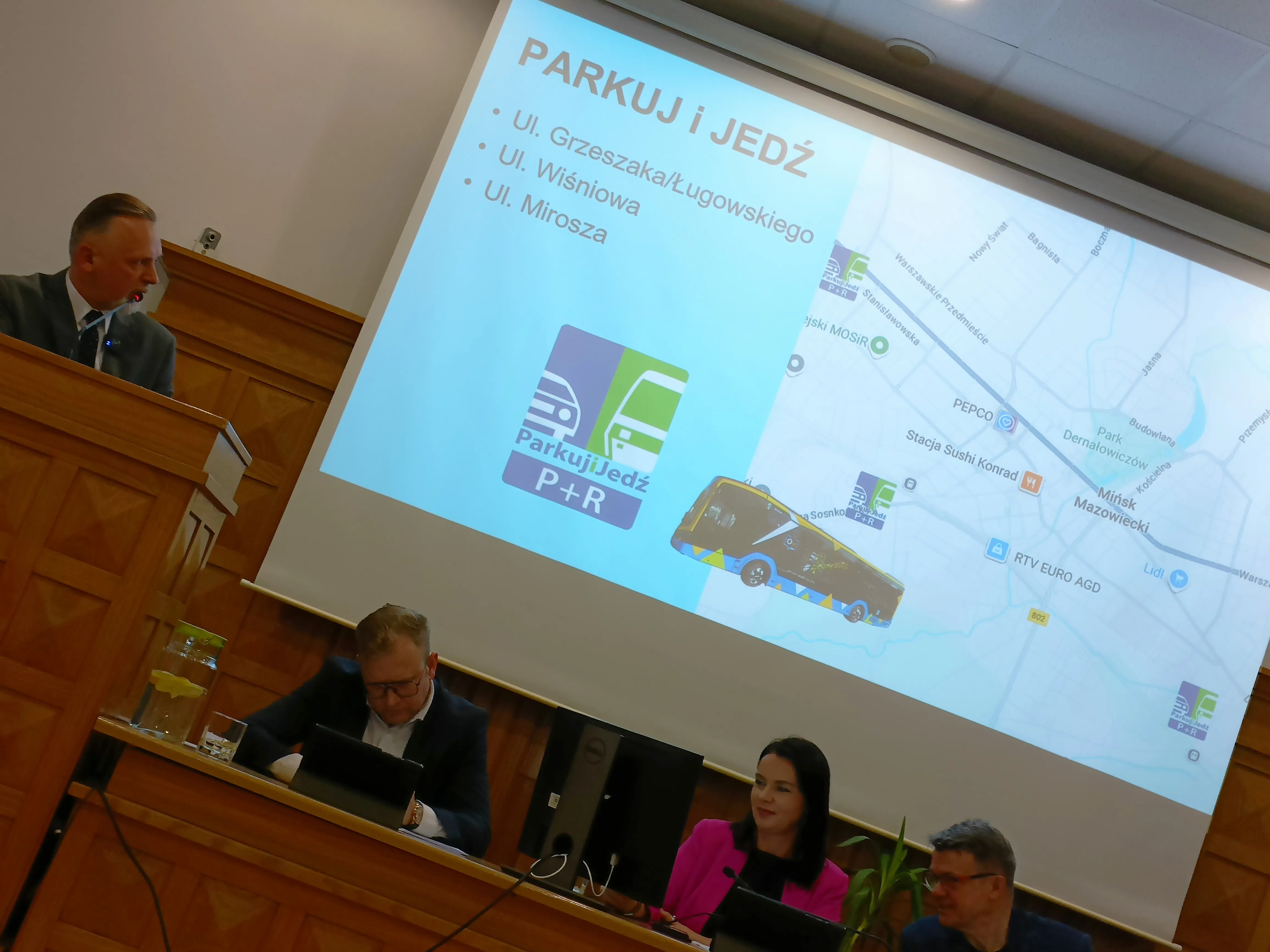 Czterech dorosłych siedzi przy stołach konferencyjnych w sali z drewnianą boazerią. Jeden mężczyzna stoi przy mównicy, trzech innych siedzi. Za nimi ekran z prezentacją o programie "Parkuj i Jedź" oraz mapą miasta.