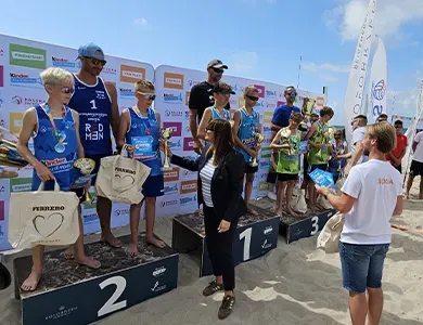 Grupa zawodników w sportowych strojach stoi na podium na plaży, odbierając nagrody od dwóch osób.