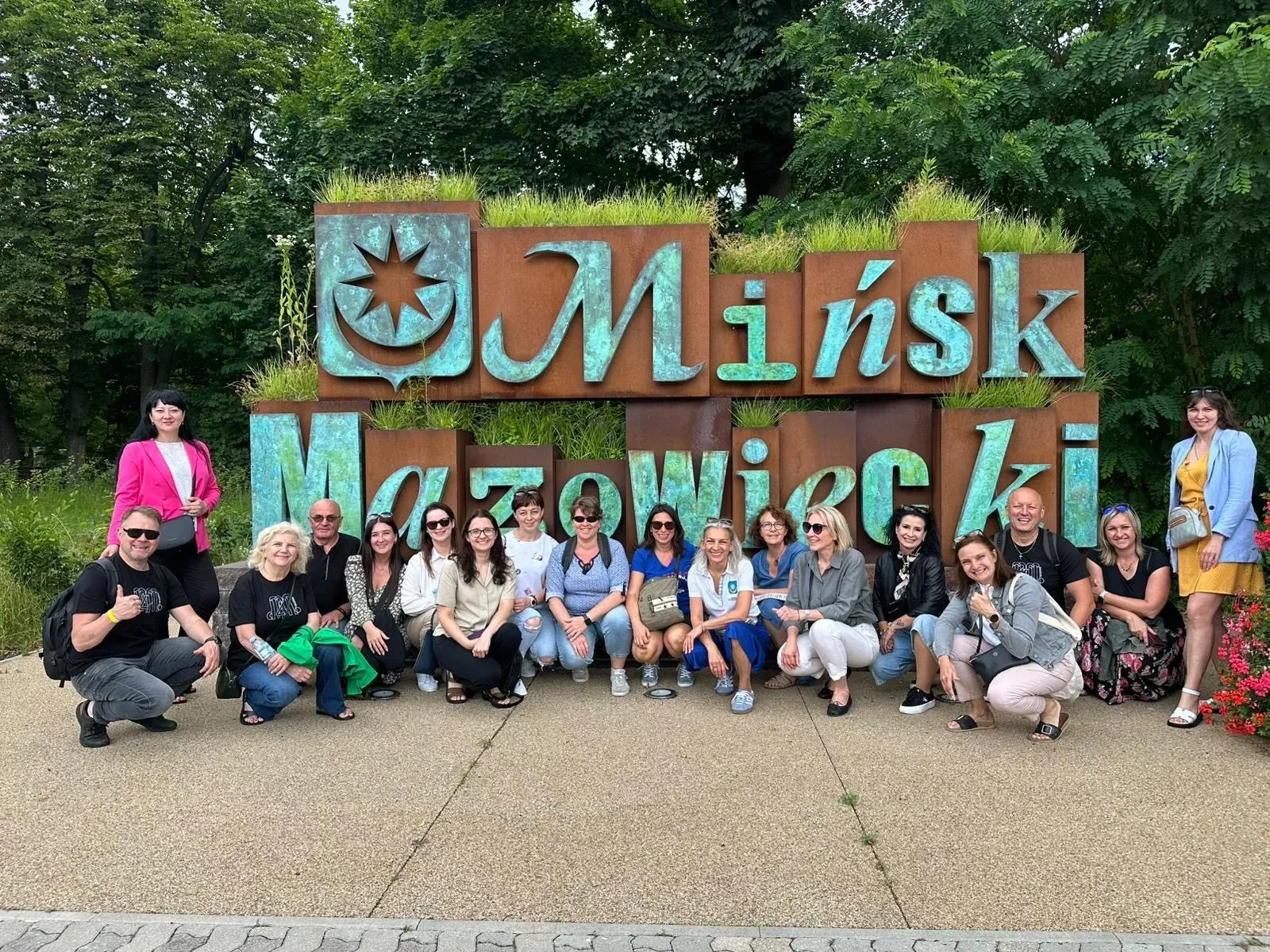 Grupa ludzi pozująca na tle dużego napisu „Mińsk Mazowiecki” z zielenią nad literami.