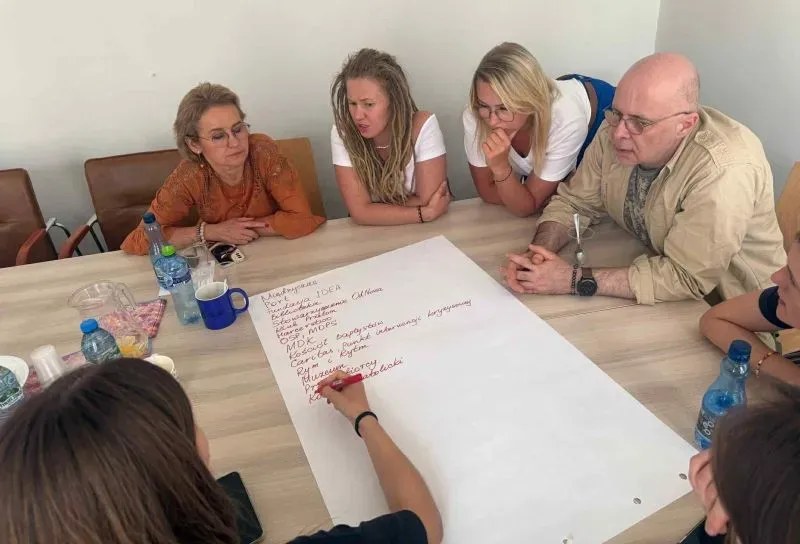 Grupa osób siedzi przy stole i wspólnie wpisuje słowa czerwonym markerem na dużej kartce.