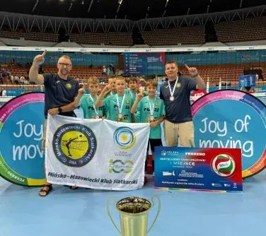 Grupa młodych siatkarzy z dwoma dorosłymi trzymająca flagę i medalami na szyi, stojąca na boisku sportowym z pucharem i dużą tablicą z napisem „Joy of moving”.