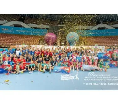 Grupa dzieci i młodzieży z trenerami pozuje na hali sportowej z medalami i pucharami, a wokół pada złoty konfetti.