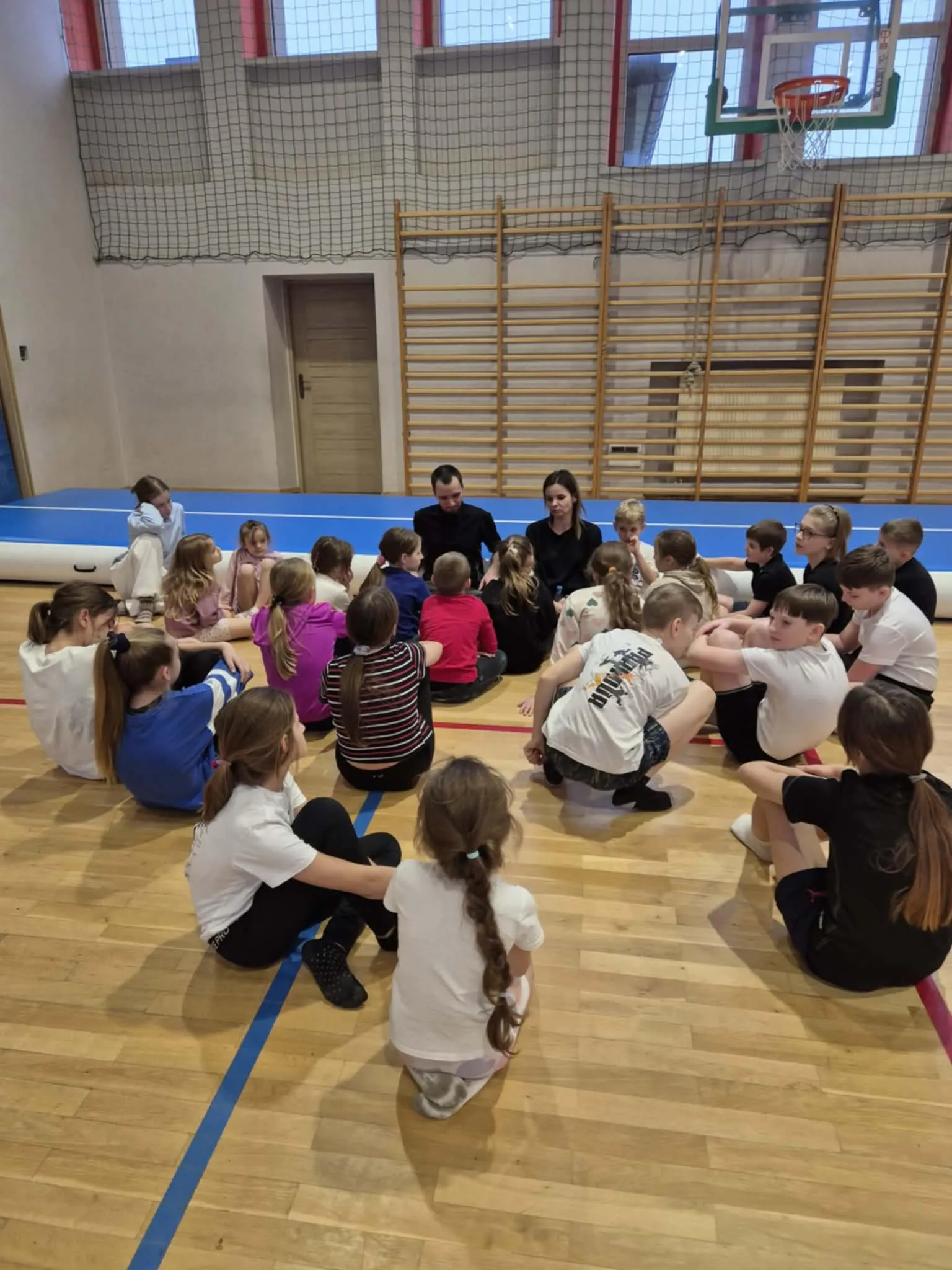 Dwadzieścioro dzieci i dwoje dorosłych siedzi w okręgu na podłodze drewnianej hali sportowej. W tle widać drabinki gimnastyczne, siatkę zabezpieczającą i kosz do koszykówki. Dzieci mają sportowe ubrania, sala jest oświetlona światłem dziennym.