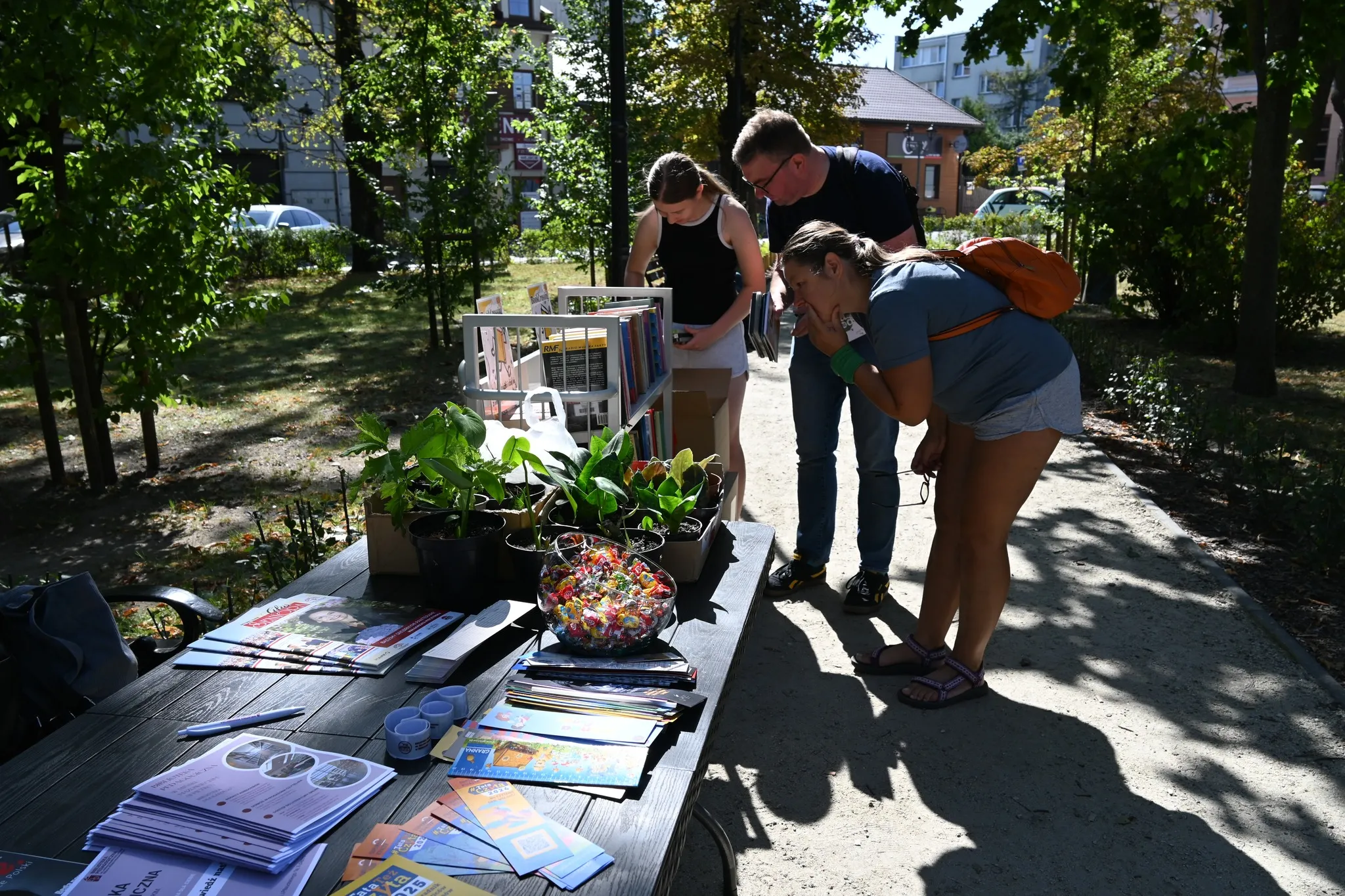 Trzy osoby stoją na zewnątrz przy drewnianym stole z książkami i roślinami, przeglądają materiały podczas słonecznego dnia w parku.