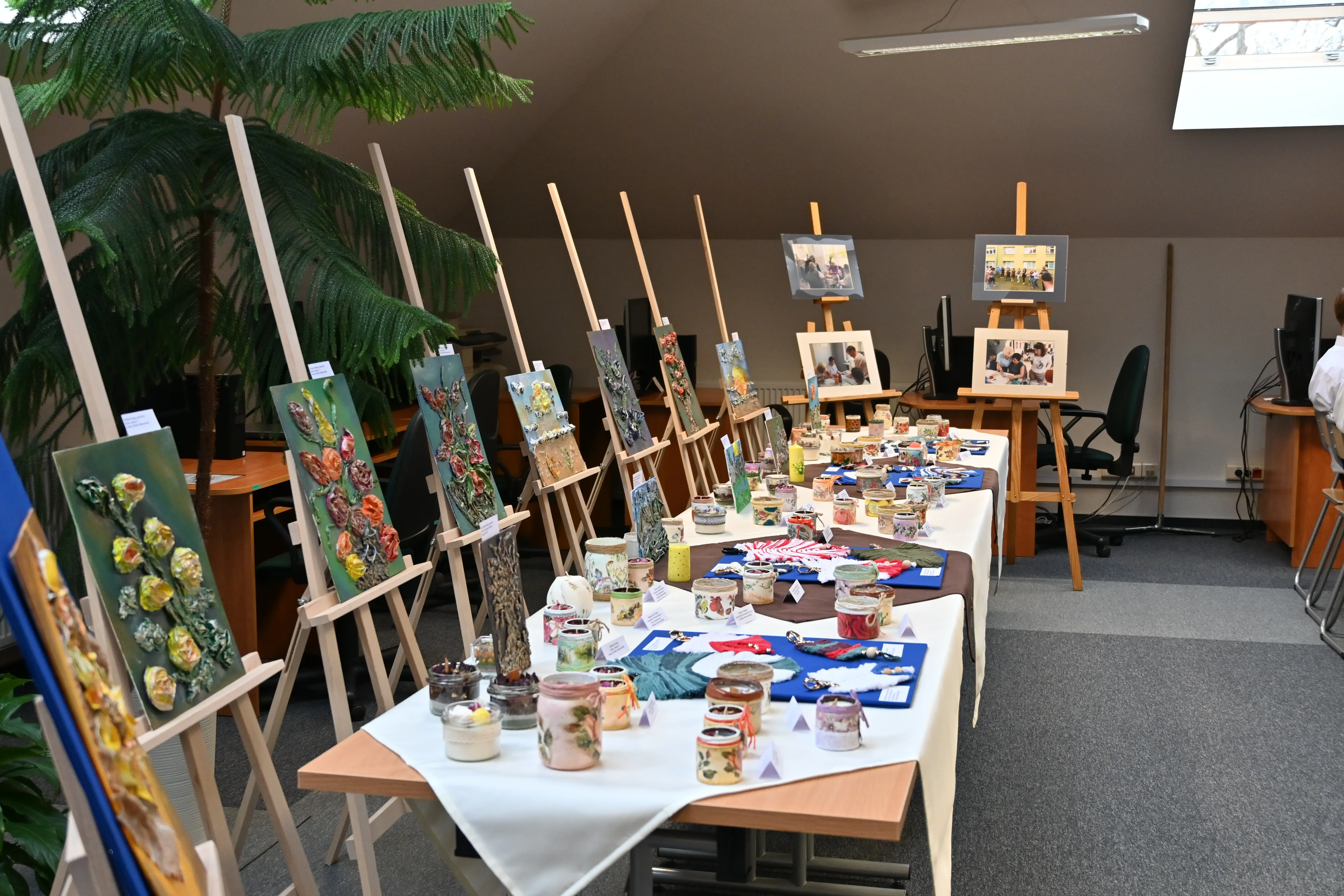Oświetlone wnętrze pokoju z wystawą prac artystycznych na stołach i sztalugach. Na sztalugach ustawiono obrazy z motywem kwiatów w relifie. Na stołach znajdują się liczne rękodzielnicze ozdoby, w tym zdobione słoiki i płaskie prace artystyczne. W tle biurka z komputerami oraz odwrócone do ściany krzesła. Pomieszczenie jest doświetlone światłem dziennym przez okno dachowe.