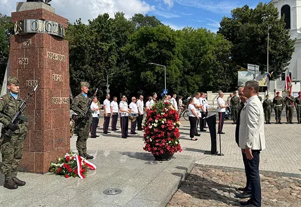 Na zewnątrz, dzień. Trzy osoby stoją przed pomnikiem z wieńcami. W tle orkiestra z instrumentami.