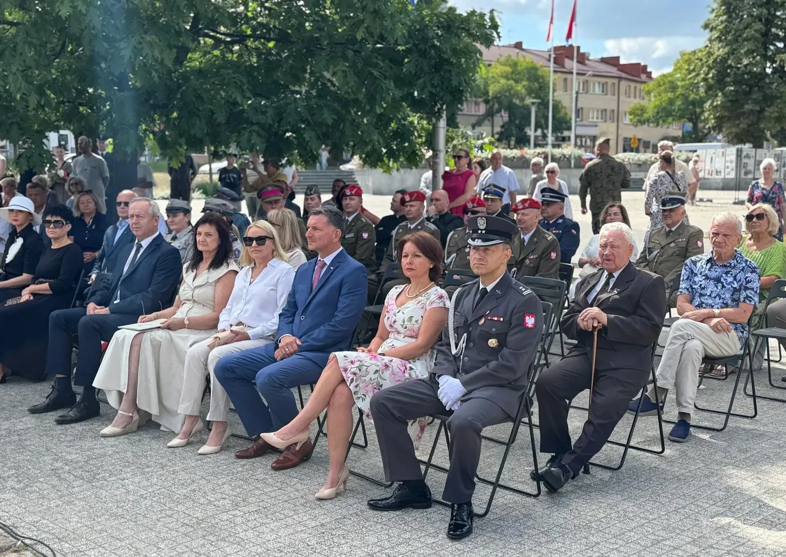 Na zawnątrz. Słoneczny dzień. Grupa około trzydziestu osób siedzi na krzesłach. Wśród nich osoby w mundurach wojskowych.