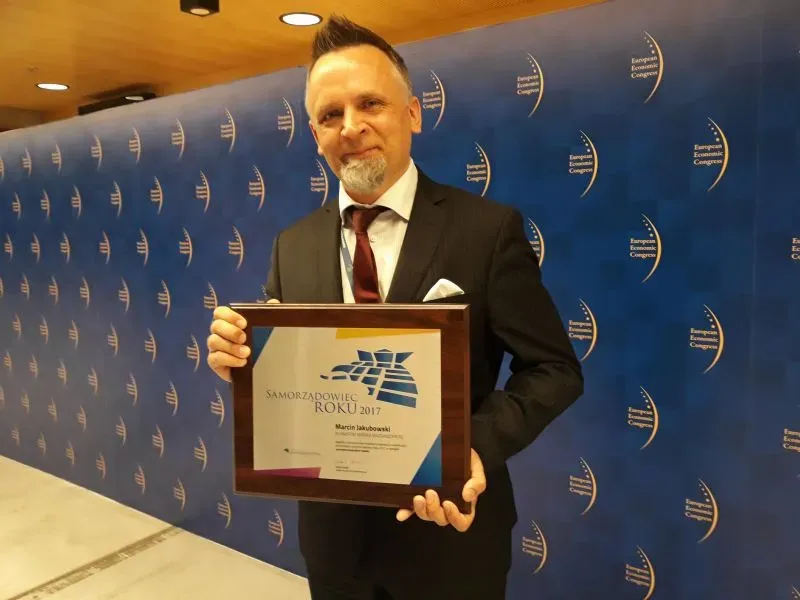 Mężczyzna w garniturze trzymający ramkę z dyplomem „Samorządowiec Roku 2017” na tle niebieskiej ściany z logo European Economic Congress.