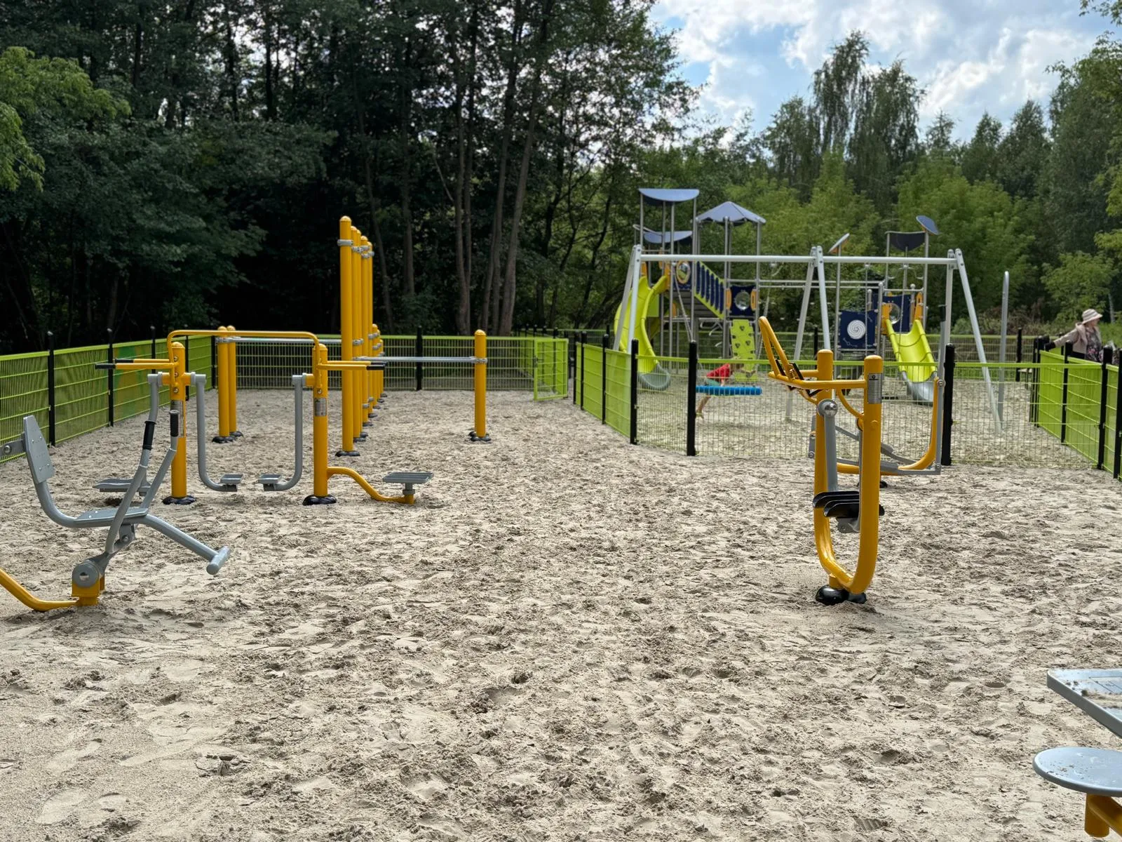 Zewnętrzna siłownia z urządzeniami do ćwiczeń na piasku obok placu zabaw z huśtawkami i zjeżdżalniami.