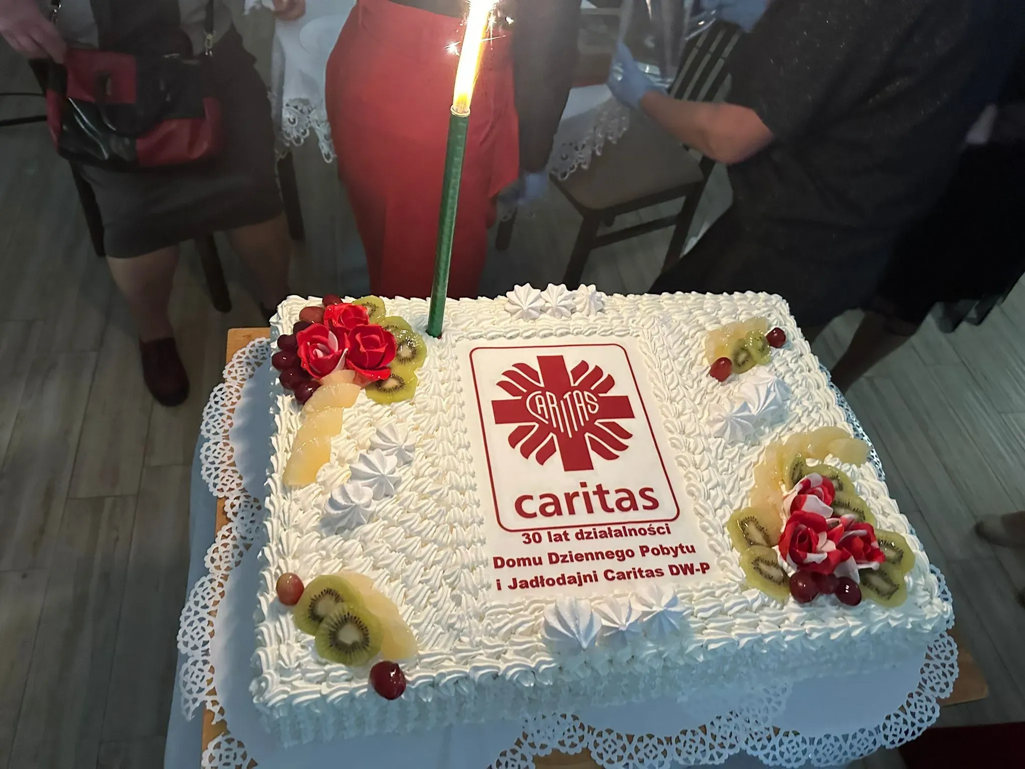 Duży prostokątny biały tort z bitą śmietaną, owocami i czerwonymi kwiatami, z zapaloną zieloną świeczką. Na środku tortu znajduje się logo Caritas i napis dotyczący 30 lat działalności. 