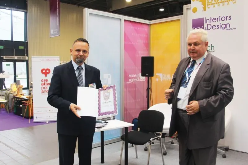 Dwóch mężczyzn w garniturach stoi w pomieszczeniu wystawienniczym, jeden z nich trzyma otwarty dokument w ramce.