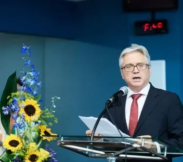 Mężczyzna w garniturze i czerwonym krawacie przemawia przy mikrofonie stojąc za mównicą obok bukietu słoneczników i niebieskich kwiatów.