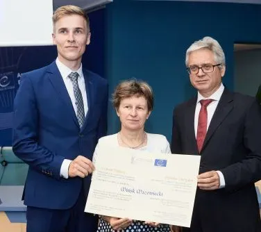 Trzy osoby w formalnych ubraniach trzymają duży certyfikat lub dokument.