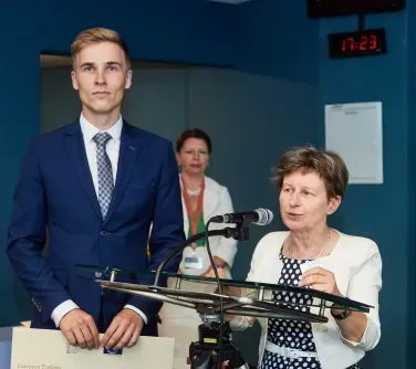 Mężczyzna w granatowym garniturze stoi obok kobiety mówiącej do mikrofonu, a w tle stoi trzecia osoba.