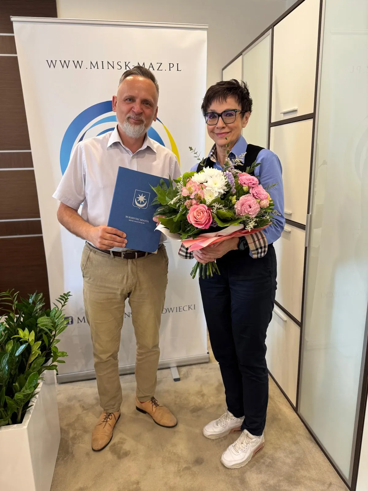 Mężczyzna trzyma folder z napisem „Burmistrz Miasta Mińsk Mazowiecki”, a kobieta bukiet kolorowych kwiatów.